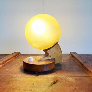 Op de afbeelding: Een tafellamp met een bolvormige, oplichtende gele glazen kap gemonteerd op een gebogen, lichtgekleurde houten steun. De voet is een ronde, donkerbruine houten schijf met een kleine witte draaischakelaar op het bovenoppervlak.