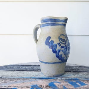 Op de afbeelding: Een vintage steengoed kan met een handvat. De gebroken witte kan heeft blauw geschilderde bloemmotieven en horizontale blauwe strepen nabij de boven- en onderkant. De kan wordt tentoongesteld op een verweerd houten oppervlak.