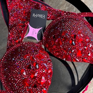 May include: A red bikini set with sparkling embellishments. The top and bottom are covered in small, iridescent crystals and larger, faceted red gems. A black tag with the text "EliteFit" is attached.