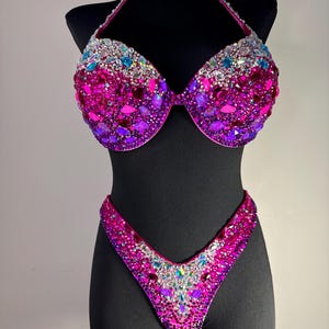 May include: A dazzling two-piece bikini set, displayed on a black mannequin. The halter-top and bottoms are covered in a vibrant array of pink, purple, and iridescent gemstones. The design is symmetrical, with a focus on the sparkling embellishments.