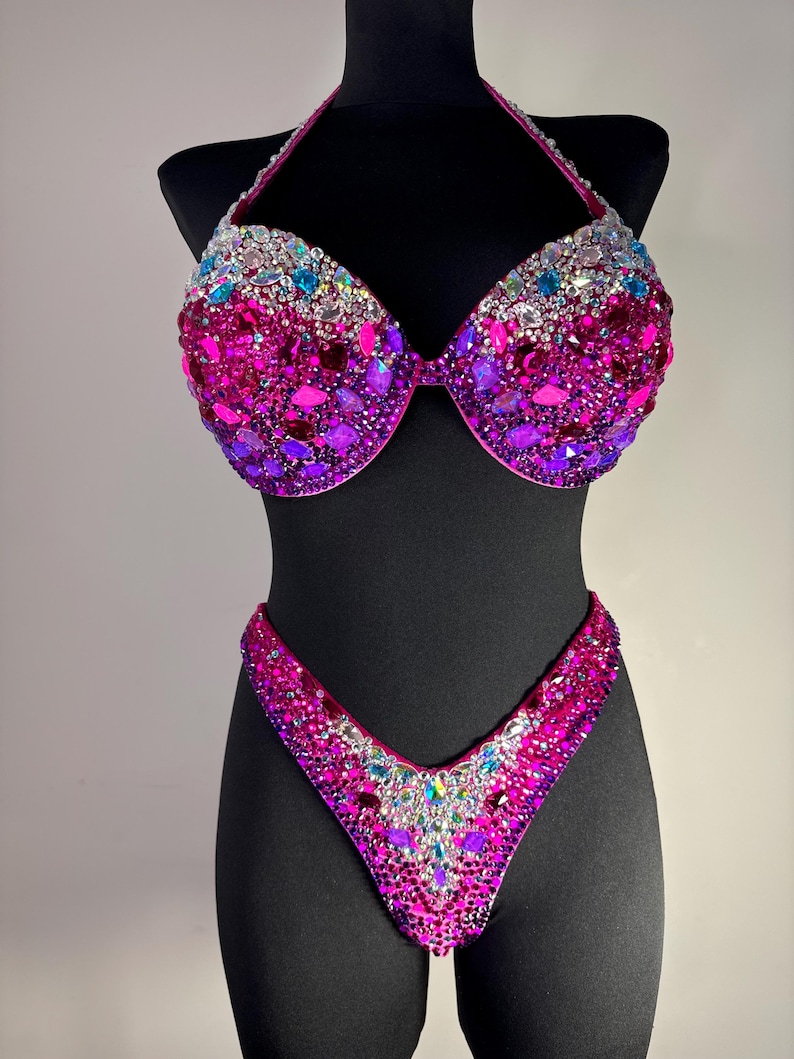 May include: A dazzling two-piece bikini set, displayed on a black mannequin. The halter-top and bottoms are covered in a vibrant array of pink, purple, and iridescent gemstones. The design is symmetrical, with a focus on the sparkling embellishments.