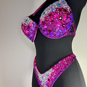 May include: A dazzling magenta bikini set, heavily embellished with a variety of sparkling rhinestones in shades of pink, purple, and blue. The bra features molded cups and the bottoms are a classic V-cut design. The bikini is displayed on a black mannequin.