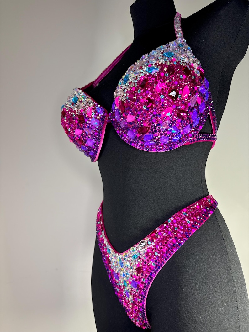 May include: A dazzling magenta bikini set, heavily embellished with a variety of sparkling rhinestones in shades of pink, purple, and blue. The bra features molded cups and the bottoms are a classic V-cut design. The bikini is displayed on a black mannequin.