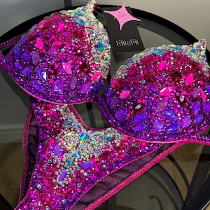 May include: A two-piece bikini set with a vibrant display of pink, purple, and iridescent gemstones. The bra and bottoms are heavily embellished with various sizes and shapes of sparkling crystals. A tag with the text "EliteFit" is attached.