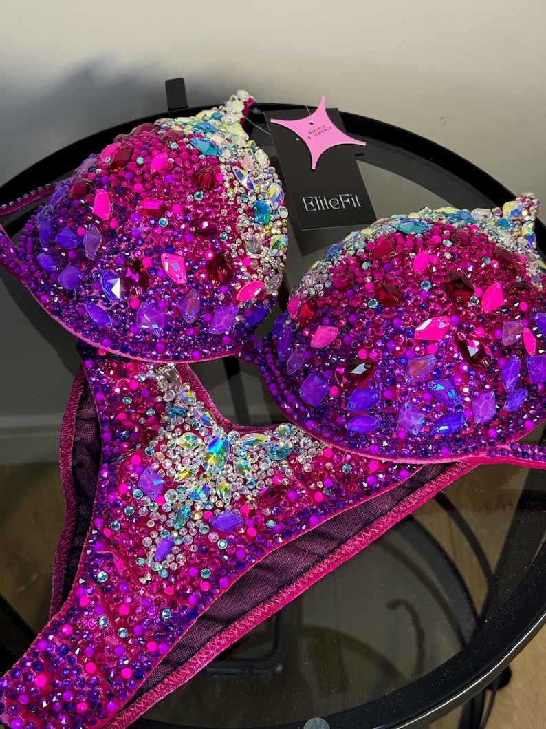 May include: A two-piece bikini set with a vibrant display of pink, purple, and iridescent gemstones. The bra and bottoms are heavily embellished with various sizes and shapes of sparkling crystals. A tag with the text "EliteFit" is attached.
