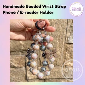 Puede incluir: Un brazalete de muñeca hecho a mano para teléfono o lector electrónico. El brazalete está hecho con cuentas iridiscentes en tonos rosa, blanco y gris. Tiene un lazo marrón y un dije plateado.