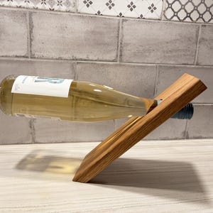 May include: A wooden wine bottle holder with a bottle of white wine. The holder is made of light brown wood and has a slanted design. The wine bottle is clear with a white label. The background is a light gray tile.