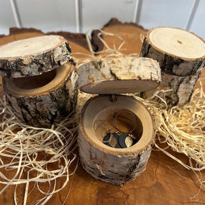 May include: Birch wood jewellery boxes, crafted from tree sections. The cylindrical boxes have lids and reveal the interior. The boxes are displayed on a wooden surface with decorative straw.