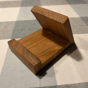 Puede incluir: Un soporte de teléfono de madera con un acabado natural. El soporte está hecho de madera maciza y presenta una base rectangular con una parte trasera elevada y un soporte inclinado. La veta de la madera es visible, lo que añade encanto rústico.