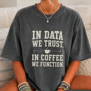 May include: A charcoal gray short-sleeve t-shirt featuring the white text IN DATA WE TRUST. IN COFFEE WE FUNCTION. with a small coffee cup graphic in the center. The shirt has a crew neckline and a relaxed fit, showcasing a vintage-style print.