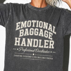 May include: A charcoal grey short-sleeve t-shirt with a vintage-style wash. The front features cream-colored text that reads: EMOTIONAL BAGGAGE HANDLER, Professional Overthinker, CARRYING EVERYONE'S FEELINGS SINCE FOREVER. The shirt has a crew neckline.