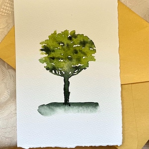 Könnte beinhalten: Ein Aquarellgemälde eines Baumes mit einer runden, grünen Baumkrone und einem dunklen Stamm. Der Baum befindet sich auf einer weißen Karte mit gezacktem Rand, die auf einem gelben Umschlag liegt. Das Kunstwerk hat einen minimalistischen Stil.