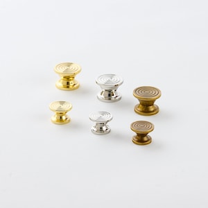 May include: Six decorative cabinet knobs in various sizes. The knobs are made of metal and have a circular, ridged design. The knobs are in shades of gold and silver.