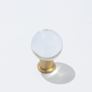May include: A clear, spherical glass cabinet knob with a brass base. The knob is a perfect sphere, reflecting light, and sits atop a short, cylindrical brass stem. The brass base has a circular design.