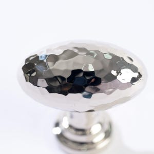 May include: A polished silver cabinet knob with a hammered texture. The oval-shaped knob sits atop a cylindrical base, reflecting light. This hardware is suitable for drawers, cabinets, and furniture.