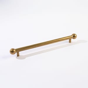 May include: A gold-toned cabinet pull with a cylindrical bar and spherical end caps. The hardware has a simple, modern design and is suitable for drawers or cabinet doors. The pull is approximately 12 inches long.