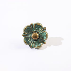 May include: A decorative flower-shaped cabinet knob with a distressed teal finish and gold accents. The knob has a central gold-colored button and detailed petal design, suitable for furniture or decor.