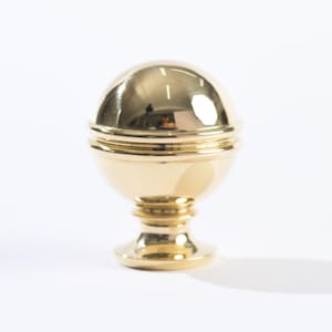 May include: A gold-toned metal finial with a rounded top and a base with three rings. The finial is a decorative element often used on furniture or drapery.