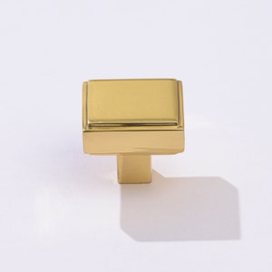 May include: A gold square cabinet knob with a raised edge and a small, square base.