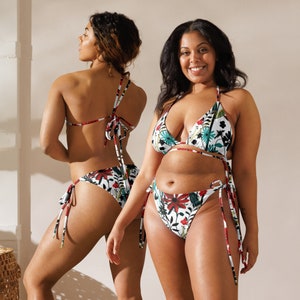 May include: Two women wearing floral print bikini tops and bottoms. The bikini tops are triangle style and the bottoms are high-waisted. The bikini has a white background with red, green, and blue floral accents.