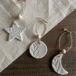 May include: Three white ceramic ornaments: a star, a circle, and a crescent moon. Each has a textured botanical design and is strung with twine and a wooden bead. The ornaments are displayed on a white cloth and a wooden surface.