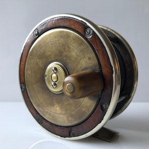 May include: A vintage brass and wood fishing reel with a wooden handle. The reel has a silver-coloured rim and a brass face with a circular design.