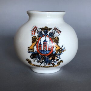 May include: White porcelain vase with a gold rim. The vase features a colorful coat of arms with lions, a castle, and flags. The text "København" is written on a banner at the bottom.