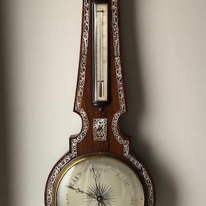 May include: Antique wooden barometer with inlaid decorative patterns. The barometer features a circular dial with a needle, a thermometer, and a smaller dial at the top. The wood is a rich brown color, and the inlays are white.