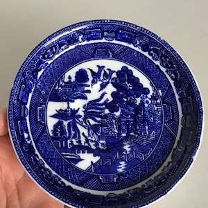 May include: A blue and white porcelain saucer with a traditional Chinese willow pattern. The saucer features a detailed design of a willow tree, a bridge, and a pagoda.