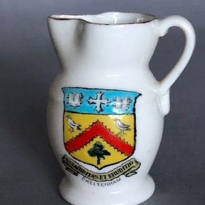 May include: A small, white ceramic pitcher with a handle and a gold rim. The pitcher features a colorful crest with a cross, birds, and a tree, along with the text "SALUBRITAS ET ERUDITIO CHELTENHAM".