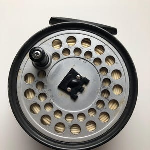 May include: A classic fly fishing reel featuring a black rim and a silver-toned spool. The spool is perforated with numerous circular holes and holds beige fishing line. A black handle and a central knob are also present.