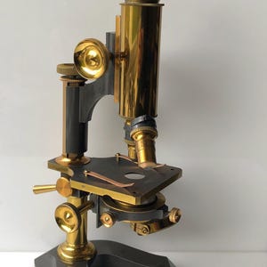 May include: An antique microscope with brass and dark gray components. The microscope features a brass eyepiece, objective lenses, and adjustment knobs. The base and supporting structure are dark gray.