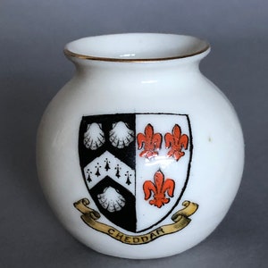 May include: A white ceramic vase with a gold rim and a black and white coat of arms with orange accents. The coat of arms features three fleur-de-lis and three scallop shells. The vase has a gold ribbon with the word "CHEDDAR" printed on it.