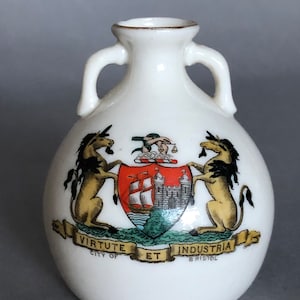 May include: White ceramic vase with two handles, featuring a coat of arms with two unicorns, a ship, and a castle. The text "VIRTUTE ET INDUSTRIA CITY OF BRISTOL" is printed on the vase.