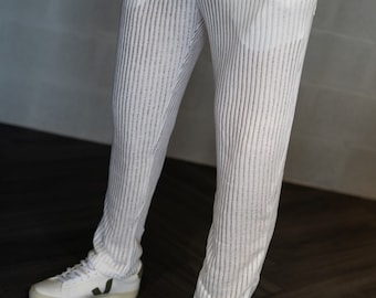 Può includere: Pantaloni bianchi caratterizzati da una texture a coste verticali e una lavorazione a maglia semi-trasparente. Questi pantaloni presentano tasche laterali e una linea dritta, abbinati a sneakers bianche con un logo verde scuro sul lato.