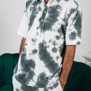 May include: A short-sleeved shirt and matching shorts set featuring a dark green and white tie-dye pattern. The shirt has a classic collar, a partial button-down placket with four light-colored buttons, and a relaxed fit made of textured fabric.