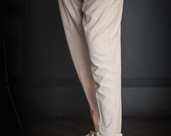 Può includere: Pantaloni beige chiaro con gamba affusolata, parte anteriore piatta e orli con risvolto, abbinati a sneakers basse color bianco sporco con finitura vissuta, suole scure e lacci chiari, indossati con una maglia bianca testurizzata a maniche lunghe.
