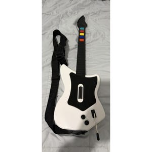 Könnte beinhalten: Ein weiß-schwarzer Videospiel-Gitarren-Controller mit einem langen Hals und fünf farbigen Griffbrett-Tasten in Grün, Rot, Gelb, Blau und Orange. Der Korpus verfügt über ein schwarzes Schlagbrett, einen Strum-Balken, zwei Drehregler und einen Whammy-Bar.
