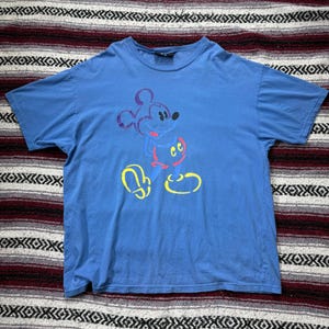 以下が含まれることがあります： ミッキーマウスのグラフィックが特徴の青いTシャツ。漫画のキャラクターは紫、赤、黄色で描かれています。このシャツは柔らかい素材でできており、半袖です。