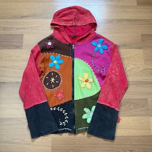 Op de afbeelding: Een patchwork hoodie met een rode capuchon en mouwen, met een verscheidenheid aan kleurrijke stoffen patches. De patches bevatten bloem- en sterapplicaties in tinten blauw, geel en bruin. De jas heeft een volledige ritssluiting en zwarte manchetten.