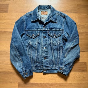 May include: A classic blue denim jacket with a button-up front, two chest pockets, and two side pockets. The jacket has a collared neckline and a small tag on the upper back. The denim has a faded, worn look.