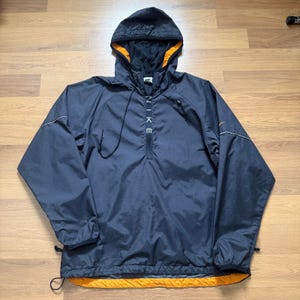 Op de afbeelding: Een marineblauwe Nike anorakjas met capuchon en halve rits. De jas heeft een zwarte capuchon met oranje voering, witte stiksels en het Nike-logo in het wit. Trekkoorden zijn aanwezig aan de zoom en manchetten.