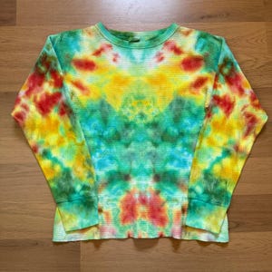 Könnte beinhalten: Ein langärmliges Thermoshirt mit einem lebendigen Batikmuster. Das Shirt hat einen Rundhalsausschnitt und Manschetten mit einer Farbpalette aus Rot, Gelb, Grün und Blau. Der Stoff hat eine Waffelstrickstruktur und das Farbmuster ist symmetrisch.