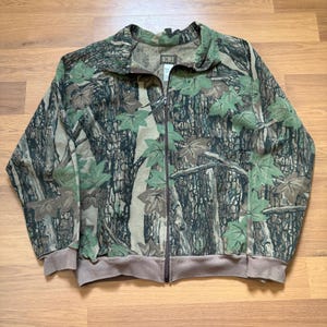 Può includere: Giacca mimetica con zip intera e colletto, polsini e orlo marroni. La giacca presenta un motivo a corteccia d'albero e foglie verdi. Il nome del marchio "OCOEE" è visibile sul colletto.