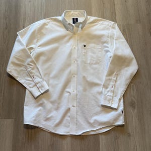 May include: A white button-down shirt with long sleeves, a chest pocket, and a button-down collar. The shirt has a small emblem on the pocket and cuff details. The shirt is made of a light-colored fabric.