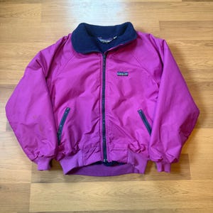 Könnte beinhalten: Eine magenta Jacke mit einem marineblauen Fleecekragen und einem durchgehenden Reißverschluss. Die Jacke hat zwei Reißverschlusstaschen und einen kleinen Logoaufnäher auf der Brust. Die Bündchen und der Bund sind gerippt.