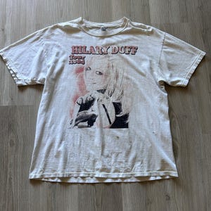 Op de afbeelding: Off-white t-shirt met een afbeelding van Hilary Duff en de tekst "HILARY DUFF Tour 2004" in roze en bruin. De shirt heeft een ronde hals en korte mouwen. Het shirt is licht versleten.