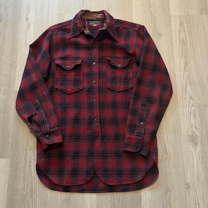 Puede incluir: Una camisa de franela a cuadros rojos y negros con dos bolsillos en el pecho y una botonadura. La camisa tiene mangas largas y un dobladillo redondeado. El cuello tiene un forro marrón.