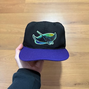 Puede incluir: Gorra de béisbol negra con visera morada. La gorra presenta un logotipo de manta raya bordado y colorido en la parte delantera. El diseño de la manta raya incluye tonos de azul, verde y amarillo. La gorra es de estilo clásico y estructurado.