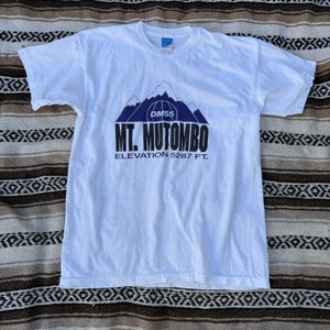 Può includere: T-shirt bianco con grafica di montagna e la scritta "MT. MUTOMBO" e "ELEVATION 5287 FT." in nero. La maglietta ha girocollo e maniche corte. L'altitudine è di circa 1611 metri. La maglietta è realizzata in un materiale morbido e confortevole.
